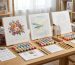 Premium wholesale paint by numbers kits displayed on a wooden studio table with pre-printed numbered canvases, acrylic paint pots, brushes, and custom retail packaging boxes.